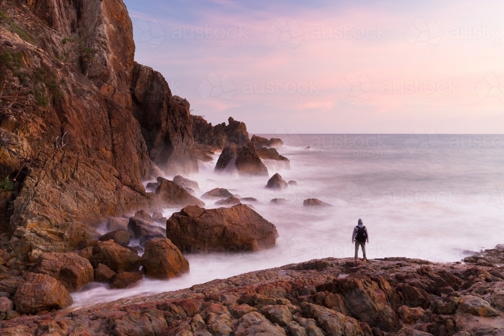 A person is silhouetted by a pink sunrise at the base of a coastal cliff in a beautiful seascape. - Australian Stock Image