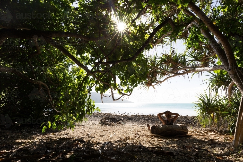 A person relaxing in the shade of tropical trees on a bright, sunny day at a remote beach - Australian Stock Image