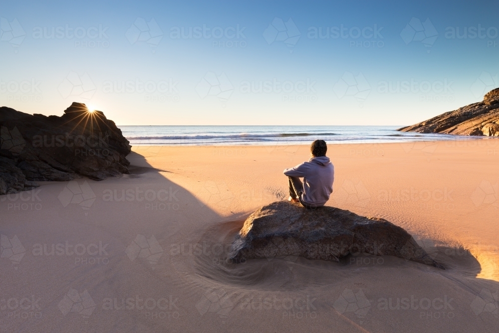 A person sits on a beautiful remote beach in Australia and watches a crisp sunrise over the ocean. - Australian Stock Image