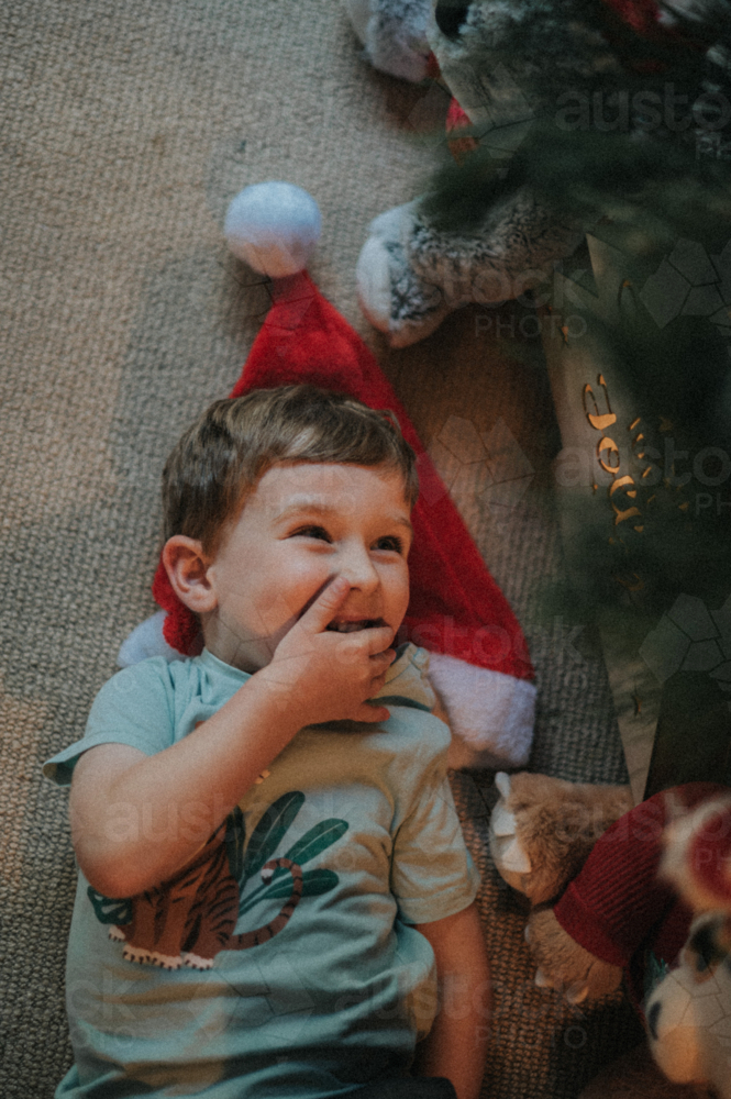 A playful child with a Santa hat laughs while lying on the floor - Australian Stock Image