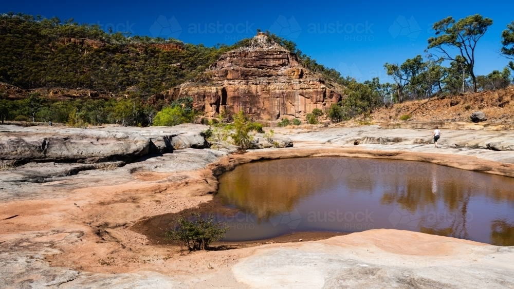 A rockpool with reflections in a starkly beautiful sandstone gorge and hiker in the distance - Australian Stock Image