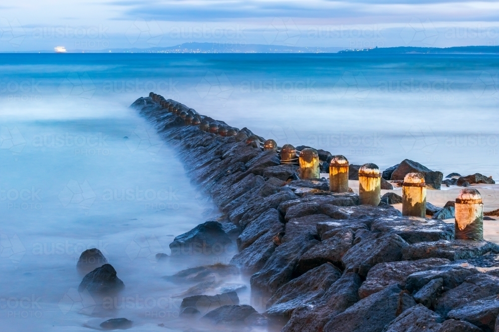 A rocky breakwater jutting out into crashing waves at twilight - Australian Stock Image