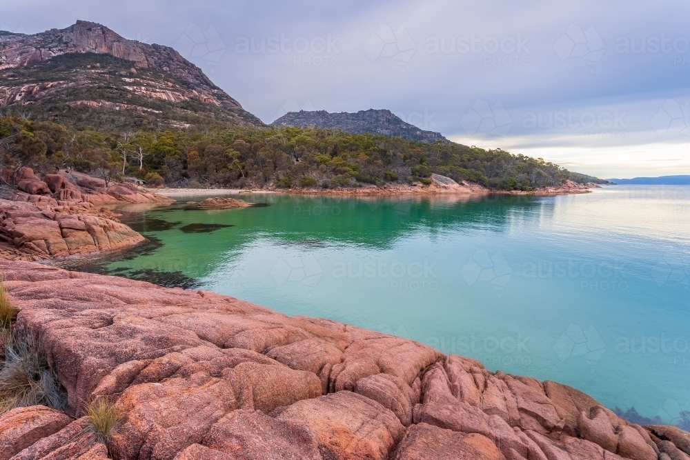 A rocky shoreline around a calm bay at the base of a mountain range - Australian Stock Image