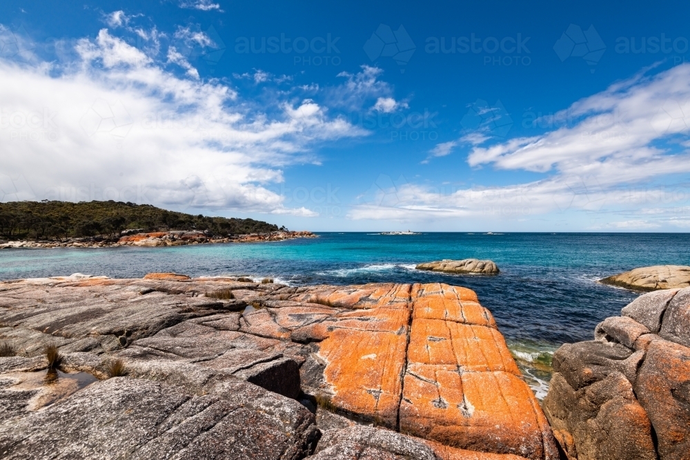 A rugged coastal scene with orange lichen covered rocks with leading lines to blue sky with clouds - Australian Stock Image