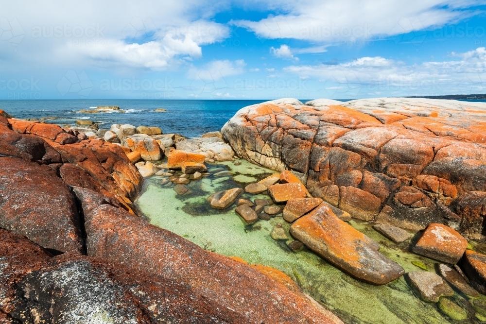 A rugged coastal scene with rock pool, orange rocks and blue cloudy sky - Australian Stock Image