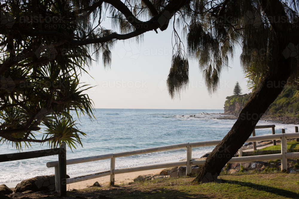 A sandy beach and grass area framed by tree foliage leads to an empty beach with a rocky point break - Australian Stock Image