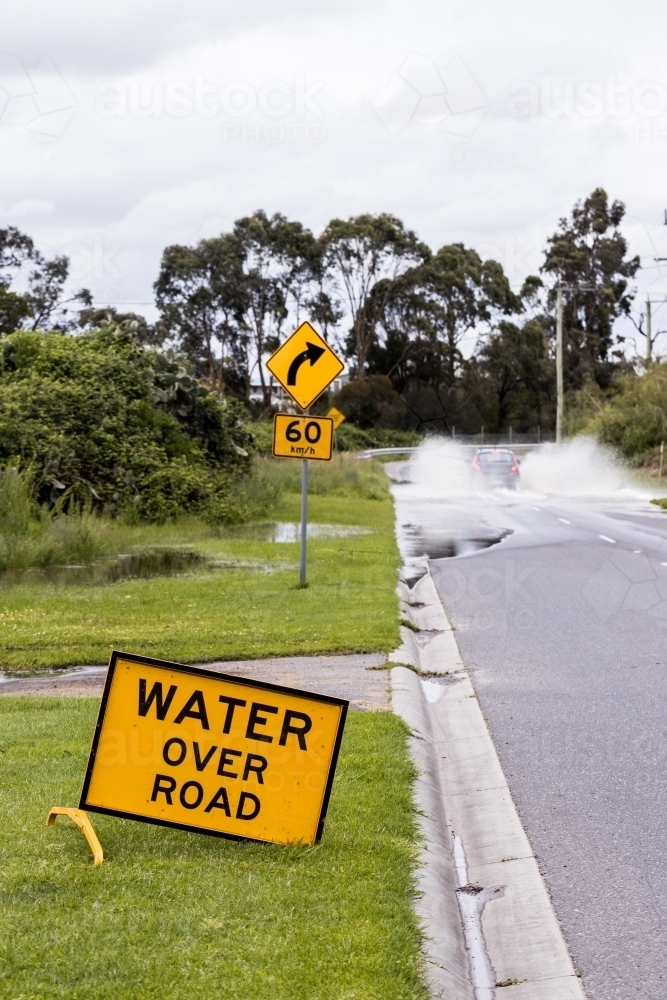 A sign which says 'Water over road' warning of flash flooding, there is a car driving through it - Australian Stock Image