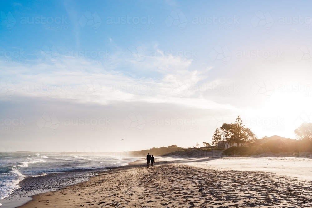 A silhouette of a couple walking closely together down the shoreline of the beach as the sunsets - Australian Stock Image
