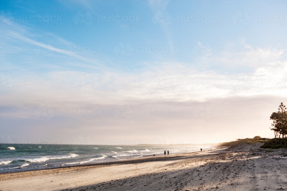 A silhouette of people in the distance walking along a beach as the sun sets. - Australian Stock Image
