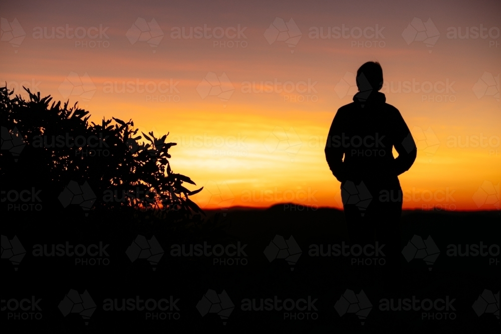 A silhouetted female stands with a vibrant coloured sunrise in front of her. - Australian Stock Image