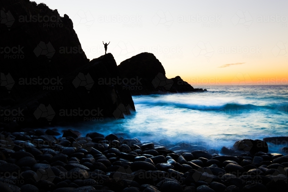 A silhouetted person welcomes the sunrise on a beach of boulders in a beautiful coastline seascape. - Australian Stock Image