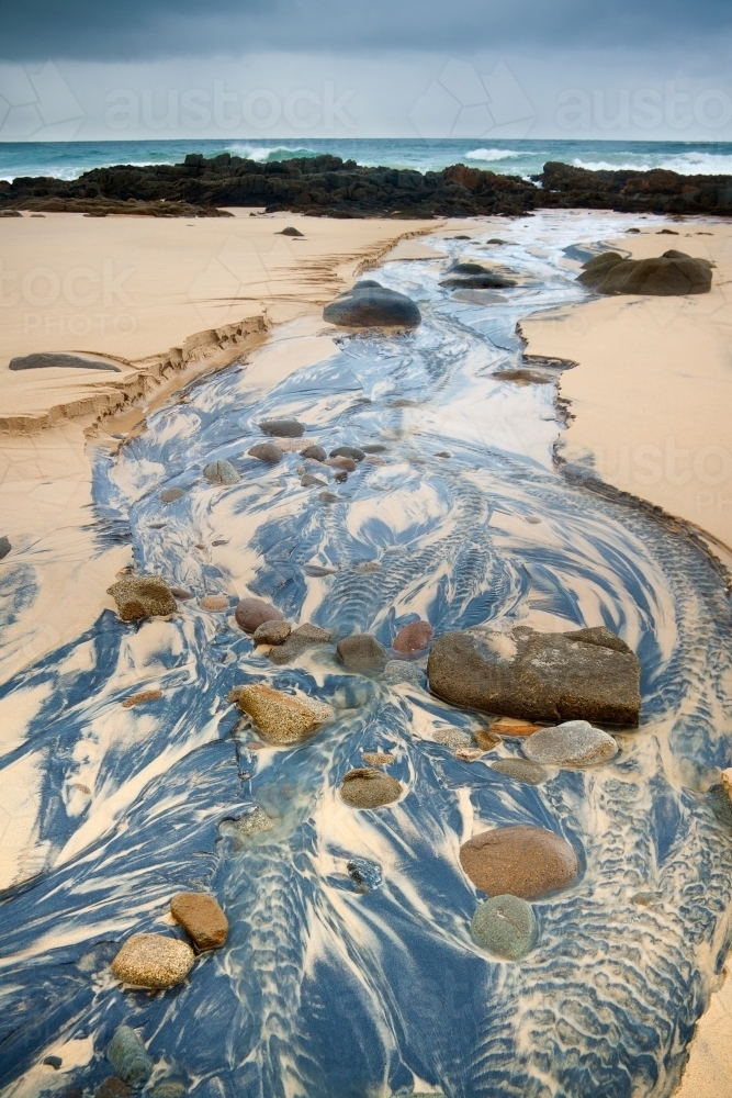 A small creek flowing down a beach creating multi colored patterns under a dark sky - Australian Stock Image