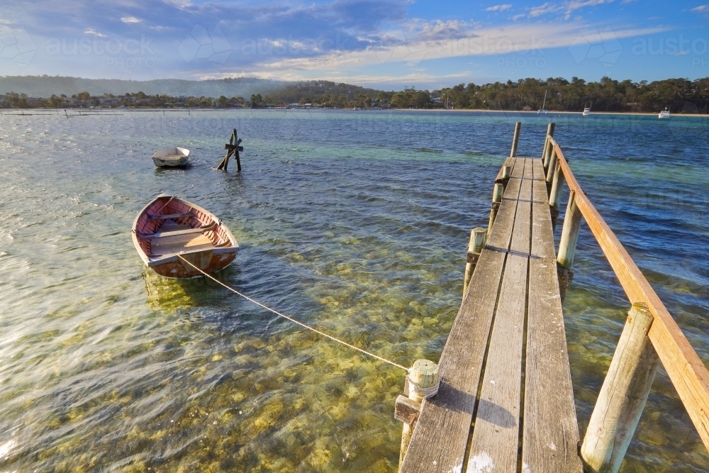 A small rowboats tied to a narrow wooden jetty over calm water - Australian Stock Image