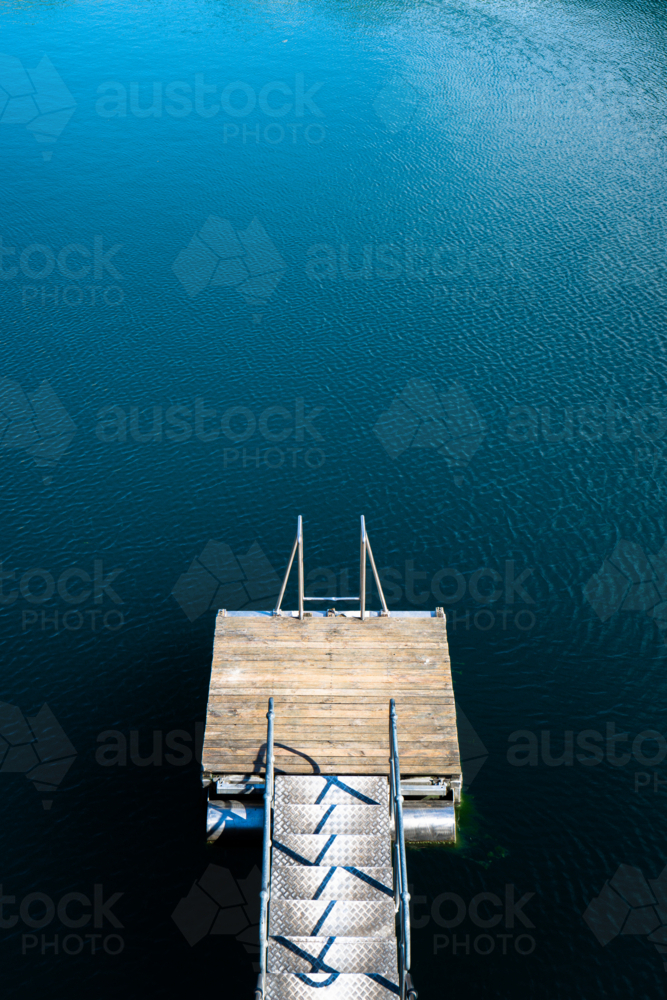 A small wooden dock extends into calm, deep blue lake water - Australian Stock Image