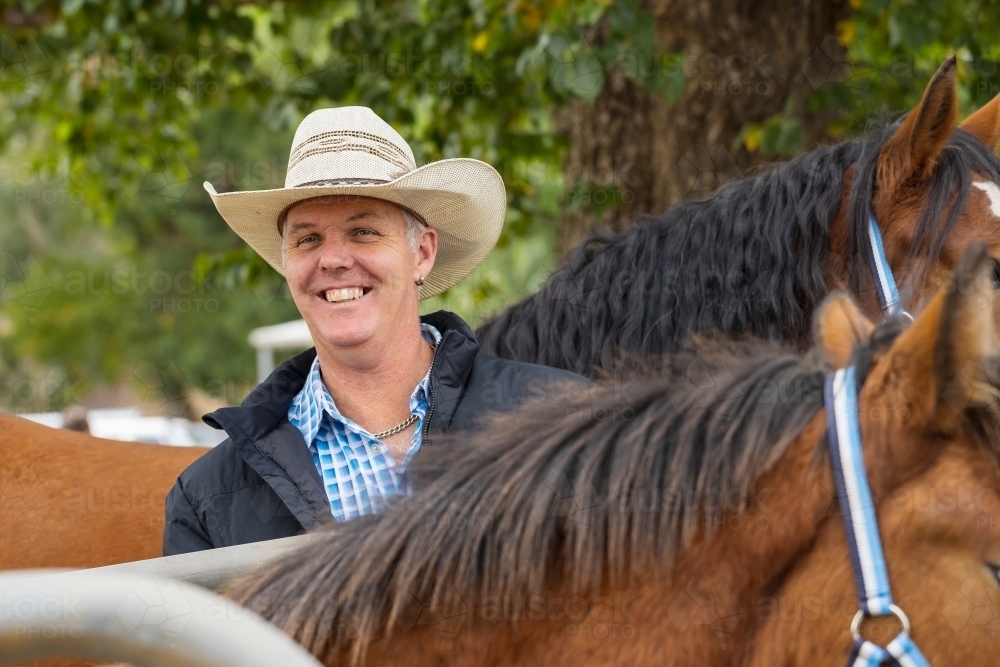 A smiling horseman wearing a cowboy hat standing among his horses - Australian Stock Image