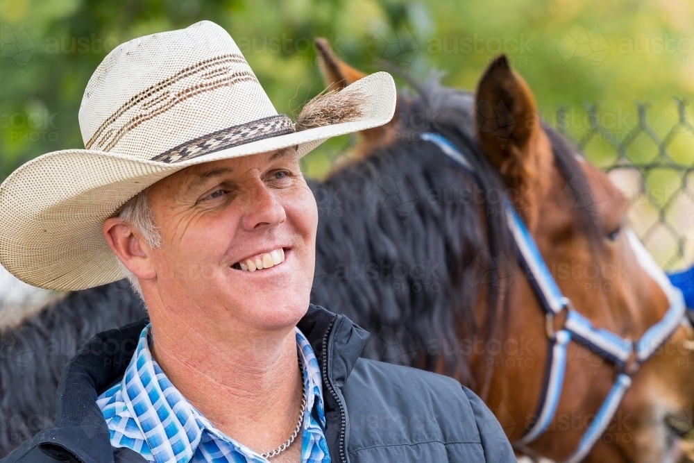 A smiling horseman wearing a cowboy hat standing among his horses - Australian Stock Image