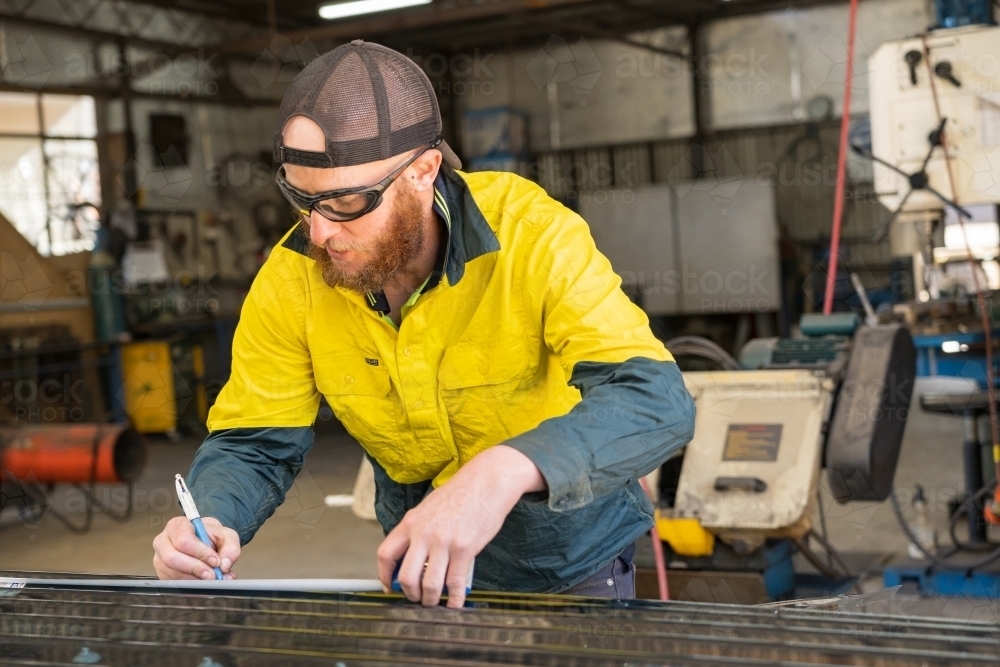 A smiling tradesman wearing high vis clothing measuring and marking along a work bench - Australian Stock Image