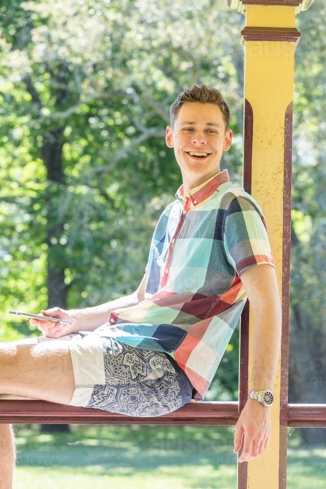 A smiling young man sitting on a wooden railing with his phone - Australian Stock Image