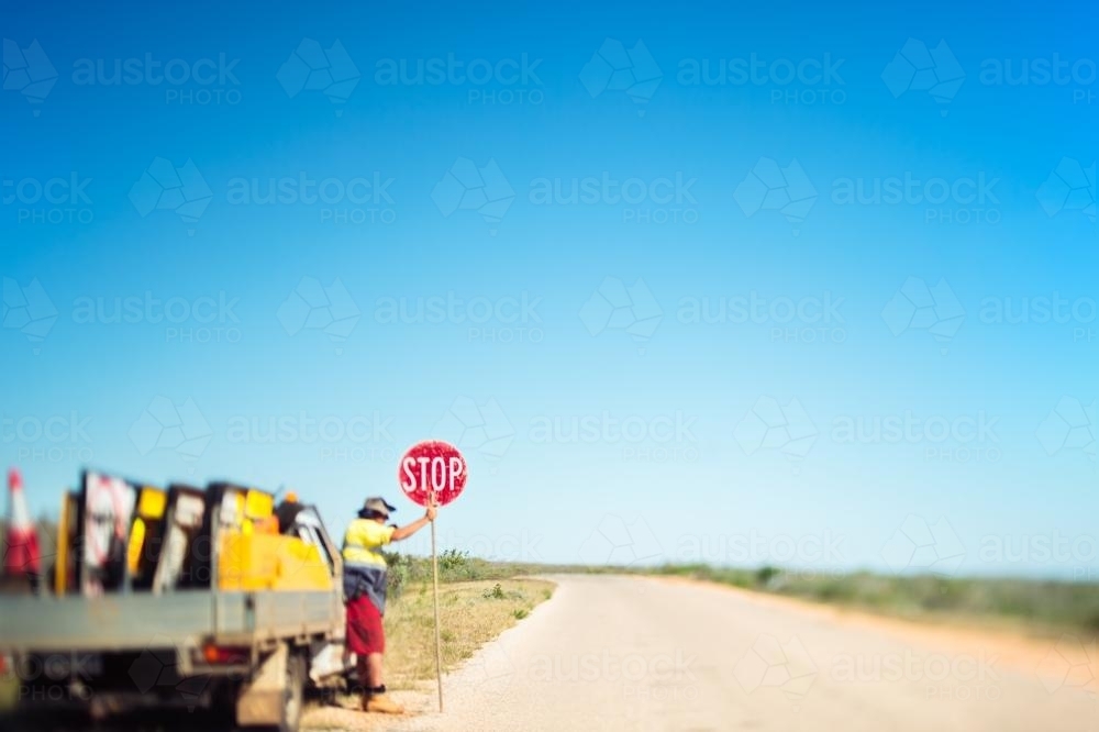 A solitary road worker holds a stop sign on a deserted road in the middle of nowhere - Australian Stock Image