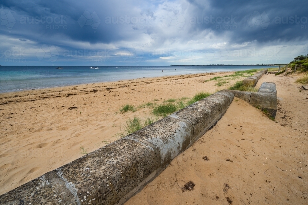 A stone retaining wall in a deep sandy beach with dark clouds overhead - Australian Stock Image