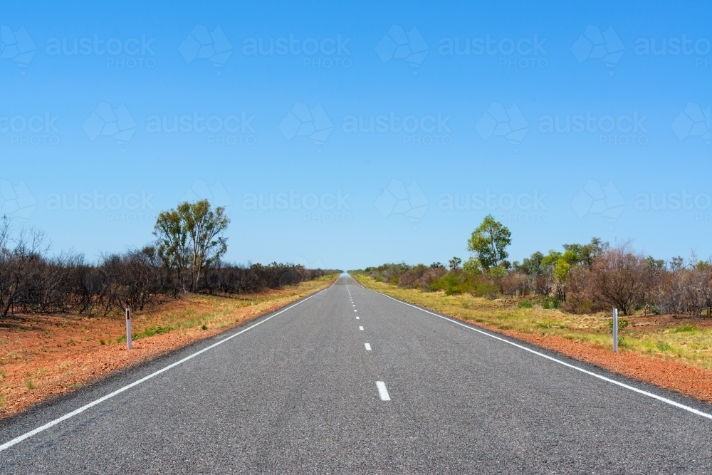 A straight bitumen road through low vegetation and orange soil with dotted white line and blue sky - Australian Stock Image