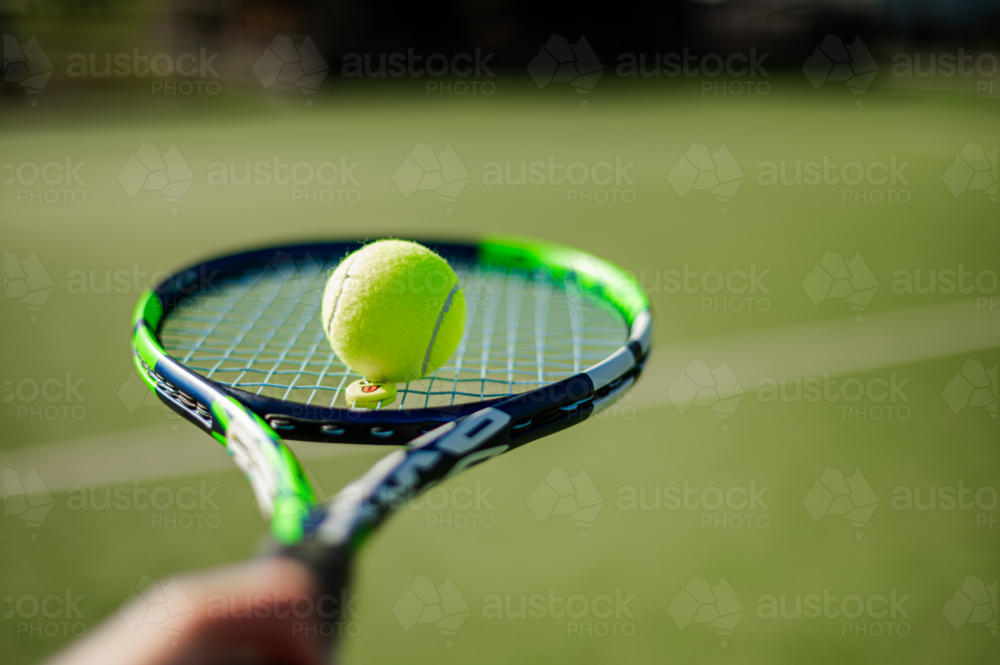 A tennis racquet balances a yellow tennis ball as the sun shines brightly on the green court - Australian Stock Image