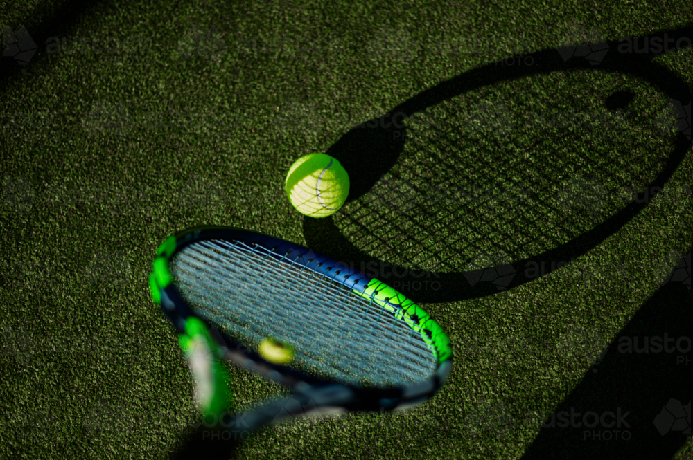 A tennis racquet hovers close to a vibrant yellow ball, both resting on lush green turf - Australian Stock Image