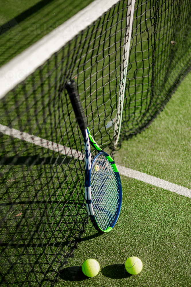 A tennis racquet leans against a net with two tennis balls on the green court surface - Australian Stock Image