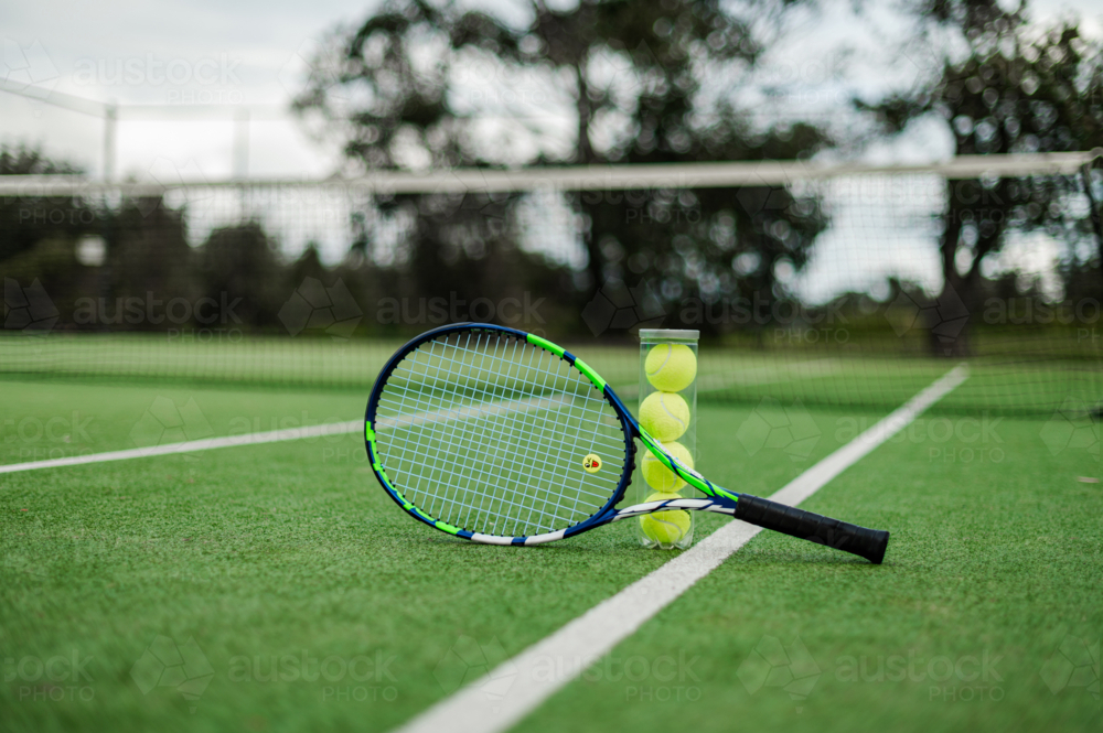 A tennis racquet propped beside a can of balls on a green outdoor court, ready for play - Australian Stock Image