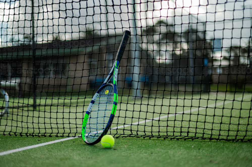 A tennis racquet rests against the net with a ball nearby on an outdoor court - Australian Stock Image
