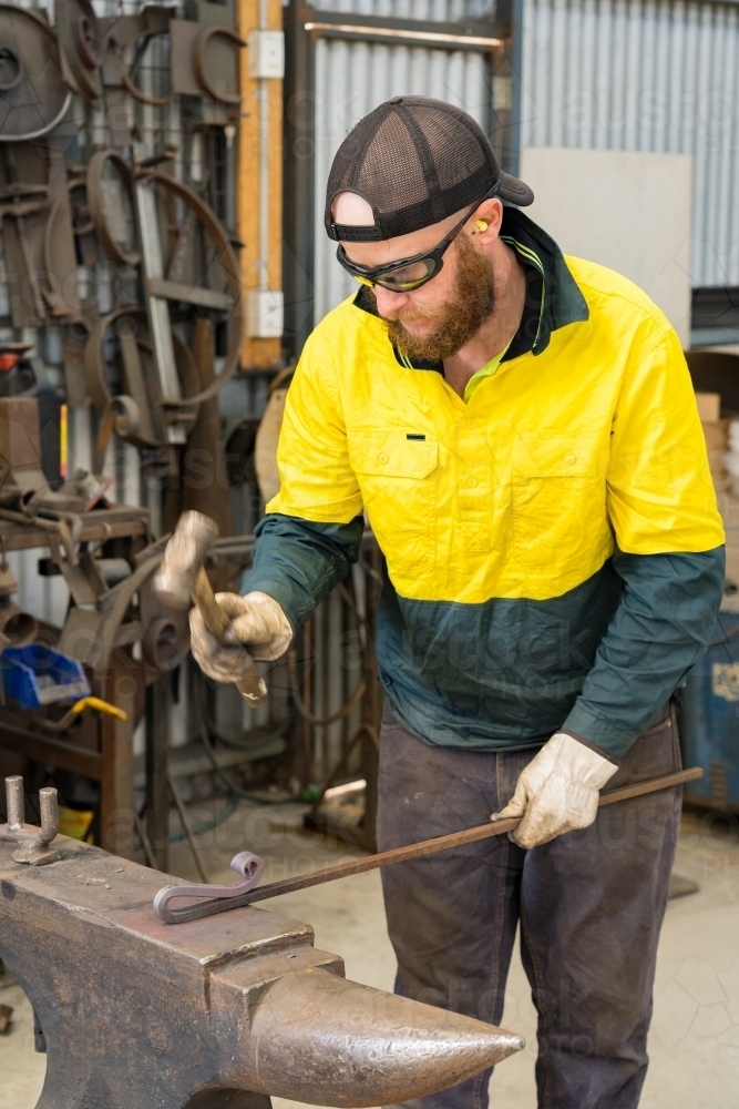 A tradesman wearing high vis clothing hammering a steel rod on an anvil. - Australian Stock Image