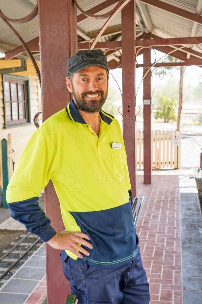 A train driver leaning against the veranda of a railway station smiling - Australian Stock Image