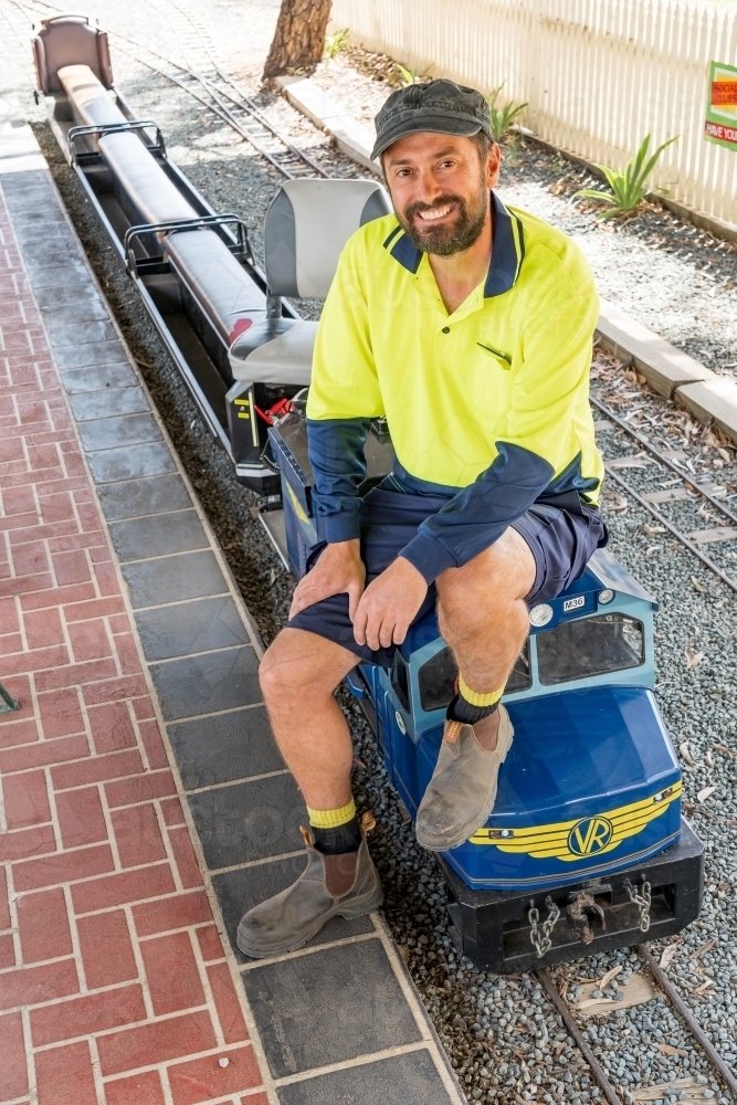 A train driver sitting on the front of a miniature railway engine smiling - Australian Stock Image