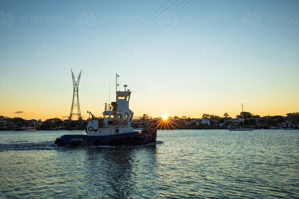 A tug boat moving upstream along the Brisbane River as the sun peaks over the horizon with clear sky - Australian Stock Image