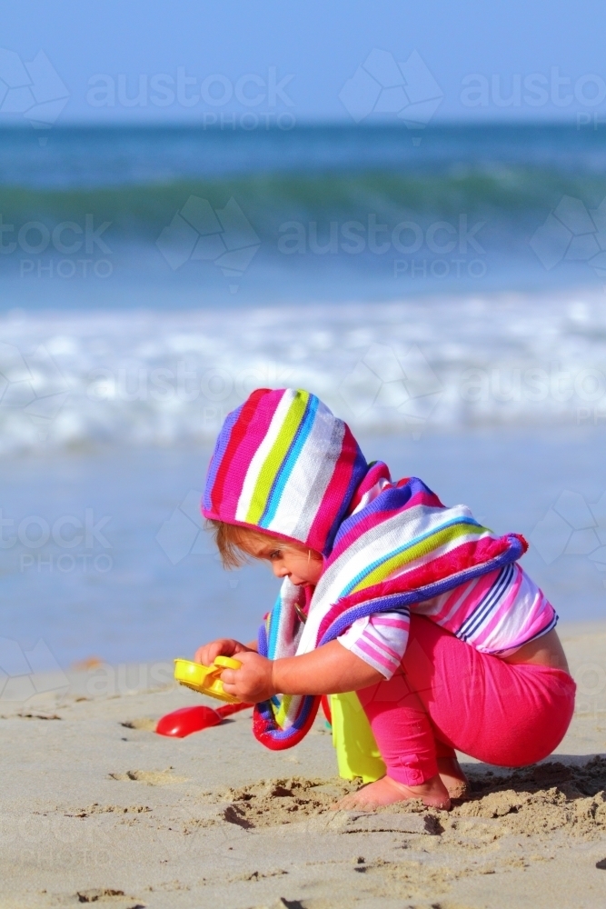 A two year old girl wearing a colourful poncho plays on a beach with her plastic toys during winter. - Australian Stock Image