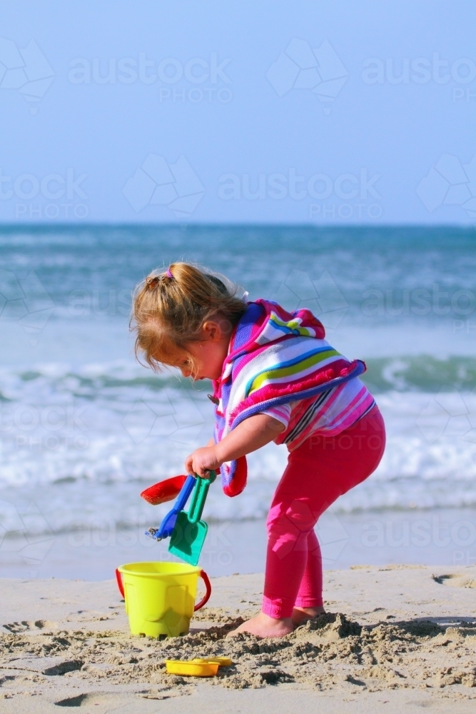 A two year old girl wearing a colourful poncho plays on a beach with her plastic toys during winter. - Australian Stock Image