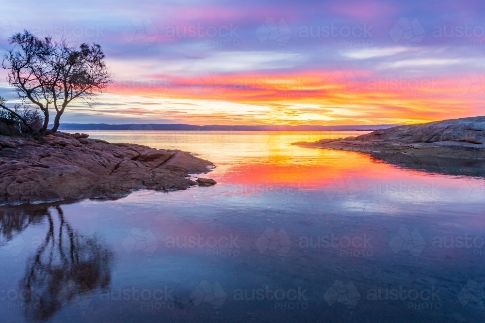 A vibrant sunset reflected in still water between two rocky points - Australian Stock Image