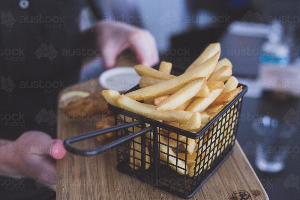 A waiter or waitress holding a basket of fried fish and chips on a wooden board in a cafe - Australian Stock Image