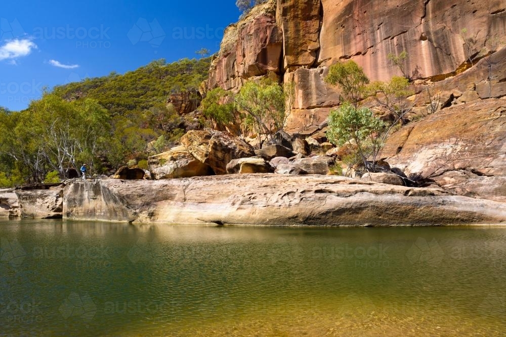 A waterhole in a gorge with orange sandstone rock formation and blue sky - Australian Stock Image