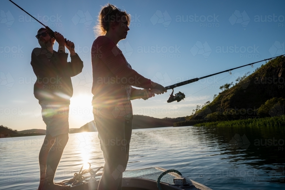 A woman and man standing and fishing from a dinghy in silhouette as sun rising - Australian Stock Image