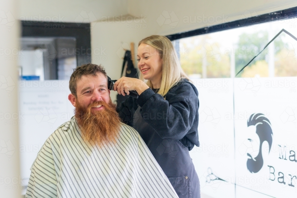 A woman barber giving a haircut to a man with a long beard - Australian Stock Image