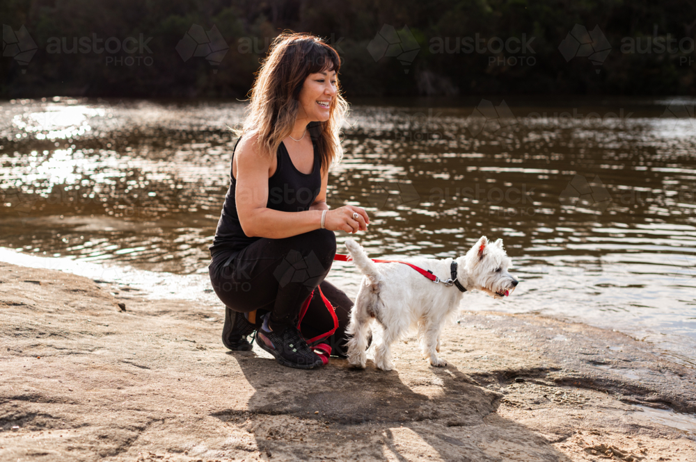 A woman crouches beside a riverbank, smiling as she interacts with her little dog near the water. - Australian Stock Image
