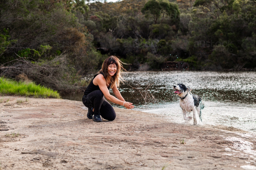 A woman enjoys a playful moment with her dog at a calm riverbank enjoying nature. - Australian Stock Image