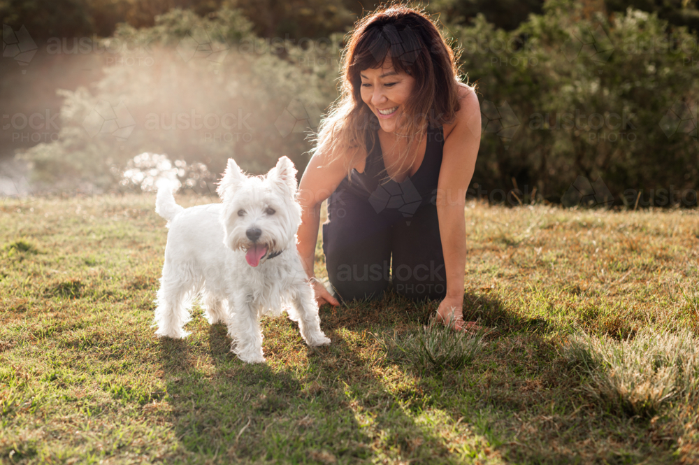 A woman enjoys time outdoors with her small white dog on a sunny day in a park - Australian Stock Image
