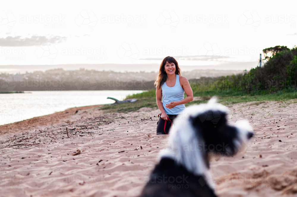 A woman happily runs with her dog on a sandy beach at sunset, enjoying the beautiful outdoor moment. - Australian Stock Image