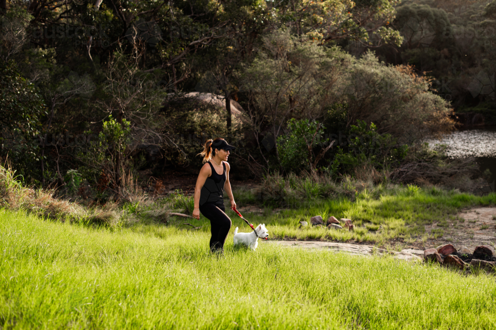 A woman in activewear guides her dog through lush grass by a serene water setting during daylight - Australian Stock Image