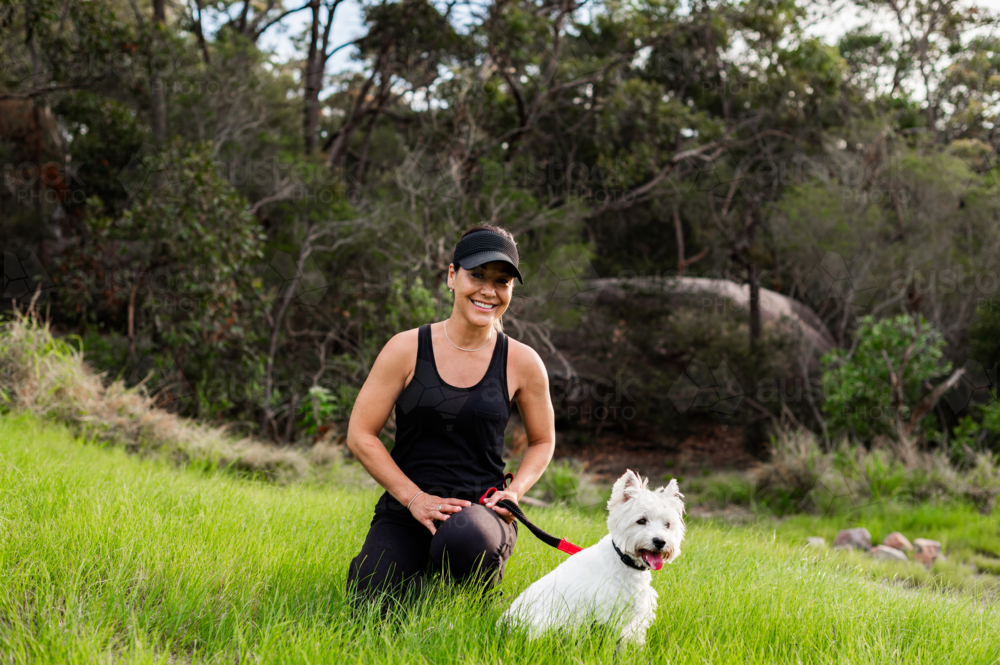 A woman kneels beside her small dog in a vibrant park, smiling contently - Australian Stock Image