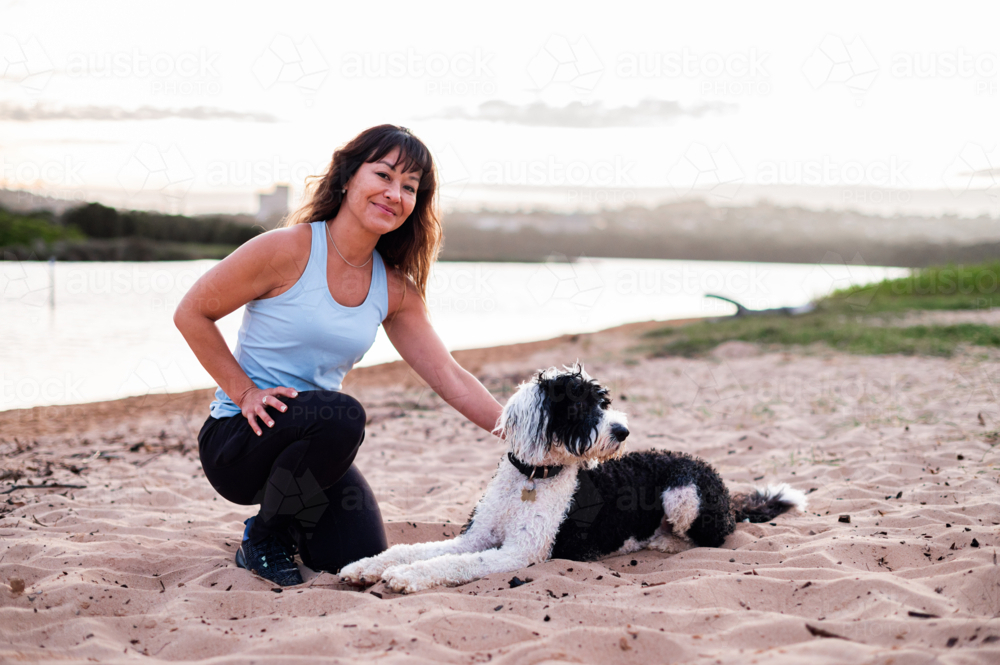 A woman kneels on the sand beside her black and white dog, smiling as the sun sets over the water - Australian Stock Image