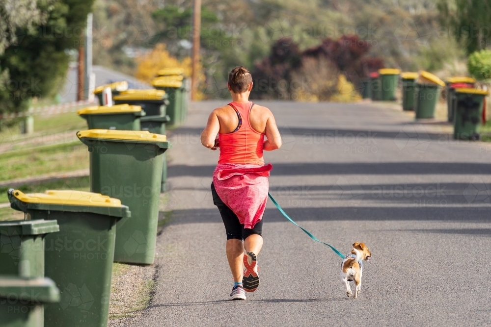 A woman running with a small dog on a neighbourhood street lined with recycle bins - Australian Stock Image