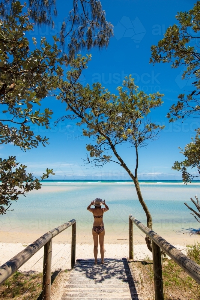 A woman stands at the end of path under a tree before a deserted sunny beach. - Australian Stock Image